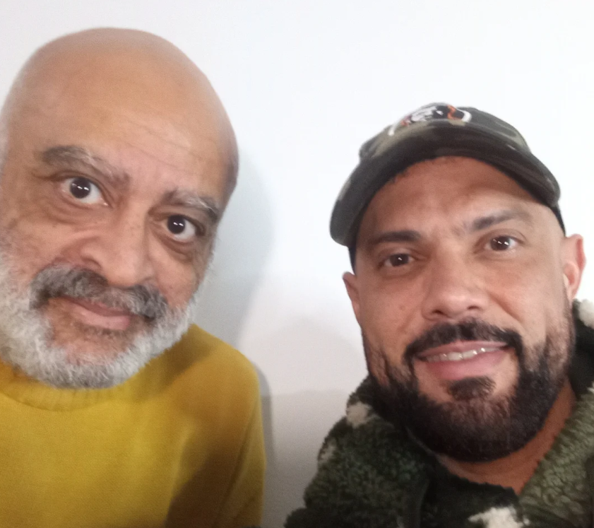 Captura de pantalla 2025-07-15 210211 Dos personas posan sonrientes tras una entrevista para el documental "Y la carne se hace verbo", centrado en diversidad e identidad. Una de ellas viste un suéter amarillo y la otra una chaqueta con gorra.