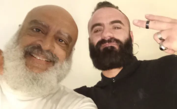 Captura de pantalla 2025-07-15 210733 Dos personas sonrientes posan tras una entrevista para un documental sobre diversidad e inclusión. Una de ellas hace el gesto de "paz" con los dedos y lleva barba espesa.