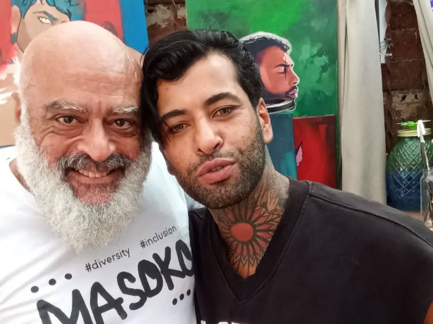Captura de pantalla 2025-07-15 211247 Dos personas posan juntas frente a obras de arte coloridas. Una de ellas lleva una camiseta con el logo de MASOKO, y la otra tiene un tatuaje visible en el cuello. Imagen tomada durante un encuentro con el artista @babylon_prince para proponerle participar en un documental.